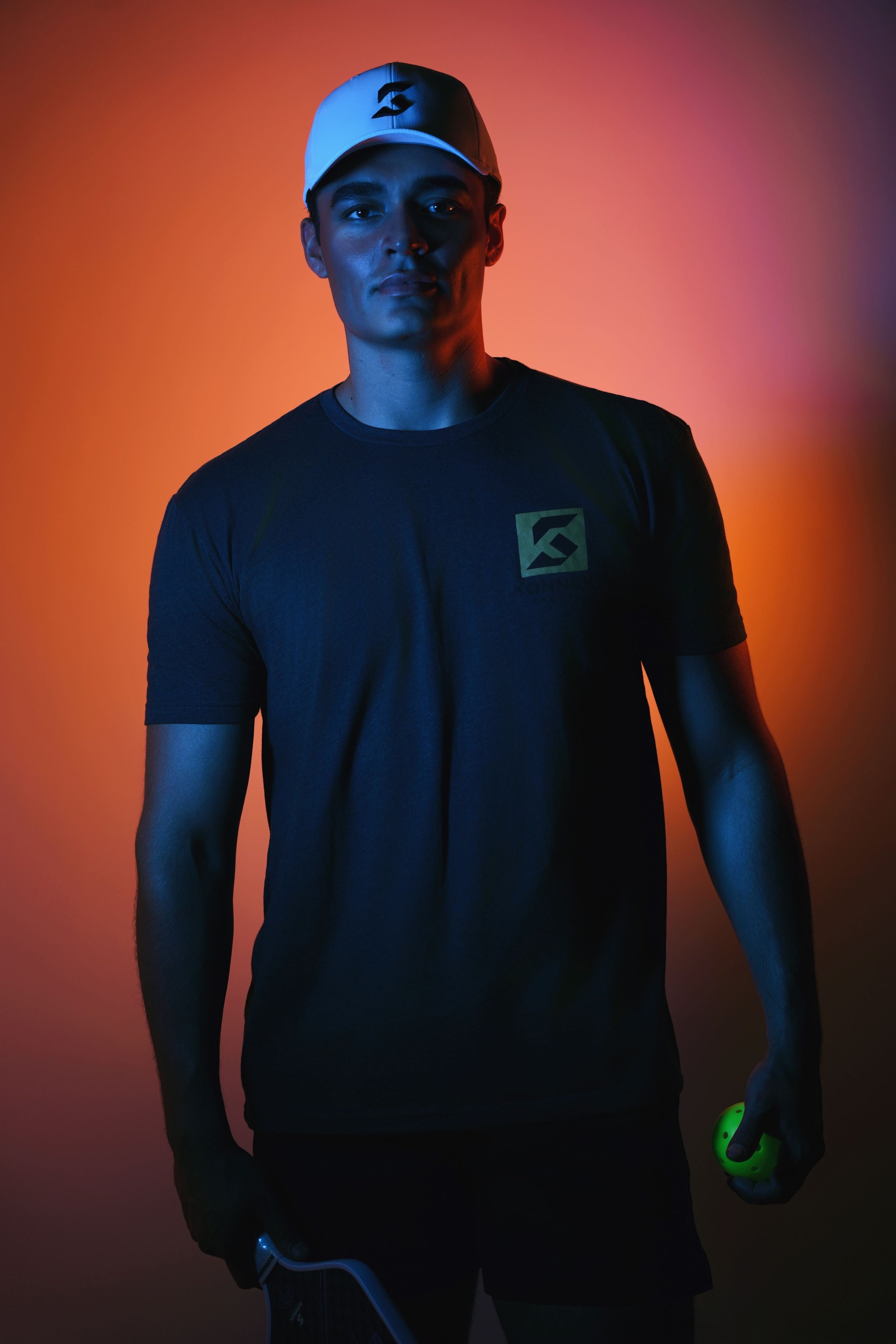 dark t-shirt with a konne ksports logo and a cap, holding a Elite Pickleball Paddle and ball